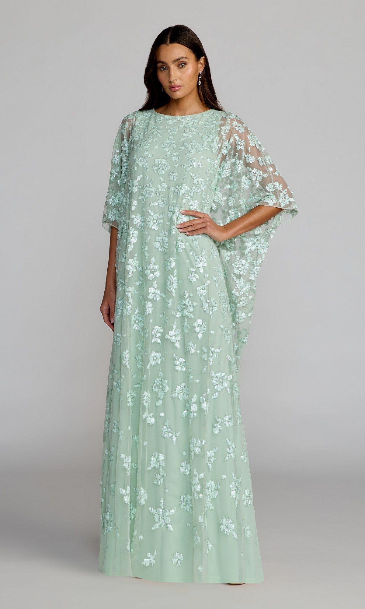 A woman wearing the Mac Duggal 9441 long formal dress in seafoamโa floor-length gown featuring floral sequins, a sheer overlay, and loose cape sleevesโstands against a plain gray background.